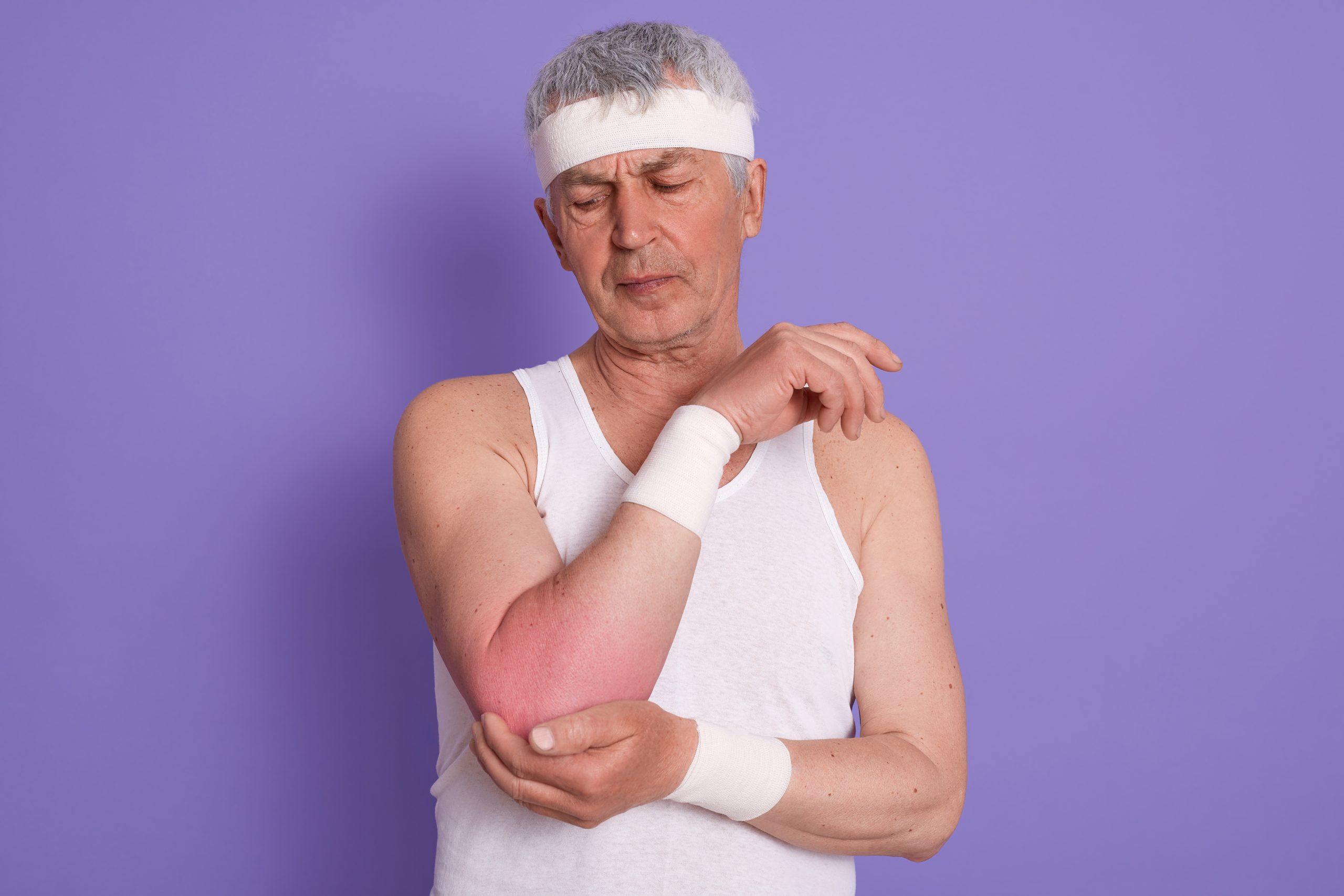 Studio shot of elderly man wearing white sleeveless t shirt and showing elbow pain.